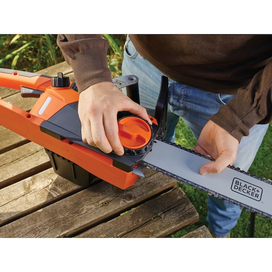 Photo of BLACK+DECKER CS2245 chainsaw, close-up of hands adjusting chain tension on wooden table outdoors.