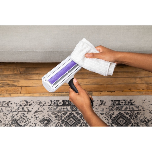 BLACK+DECKER HMSCT0001 cleaning tool with purple roller and white cloth, held above wooden floor and patterned rug.
