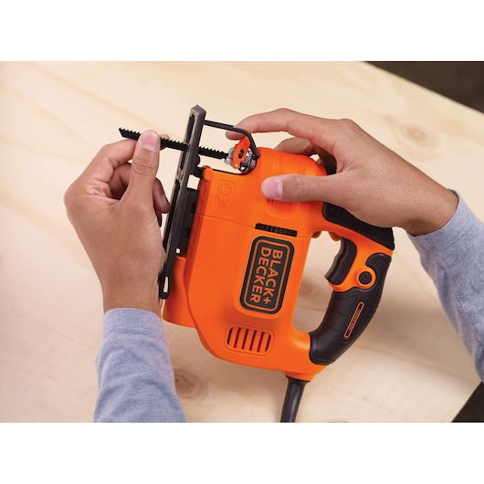 A person installing a saw blade into an orange BLACK+DECKER jigsaw on a wooden surface.
