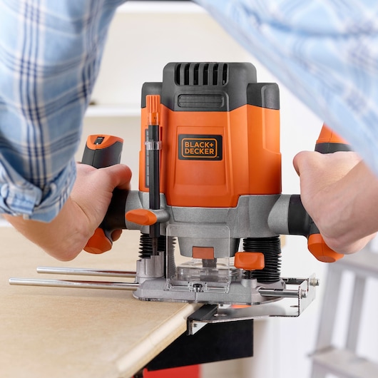 Photo of BLACK+DECKER KW1200E router held by hands while trimming wood edge, orange and black tool in use.