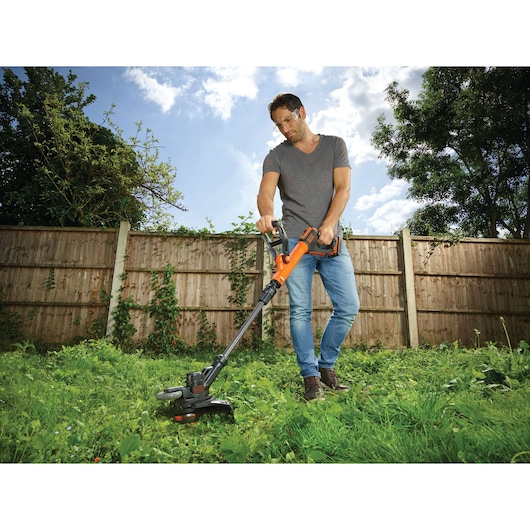 BLACKDECKER STC1820EPC grass trimmer in use outdoors, orange and black, cutting tall grass in a garden.