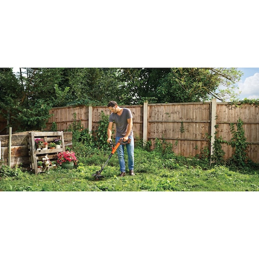 BLACKDECKER STC1840EPC cordless grass trimmer in use in a garden setting with wooden fence and plants.