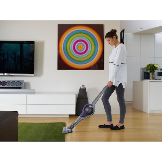 A person is vacuuming a green rug with a BLACK+DECKER cordless stick vacuum cleaner in a modern living room. The room features wooden floors, a white cabinet, a colorful circular artwork on the wall, and contemporary decor.