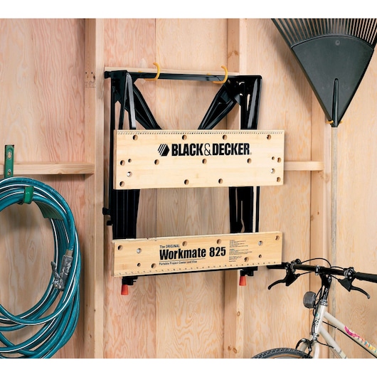BLACKDECKER WM825 folding workbench mounted on a wooden garage wall, surrounded by a hose, rake, and bicycle.